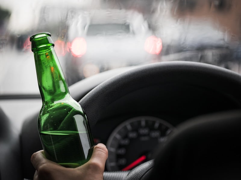 driver hand holding green beer bottle on road with light bokeh of cars
